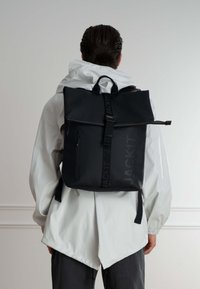 Zaino roll-top nero con finitura opaca, dotato di una tasca anteriore con zip e spallacci regolabili. Logo stampato sul lato.