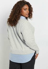 Felpa grigia chiara con polsini a costine, sovrapposta a una camicia button-up a righe blu e bianche. Morbido al tatto, vestibilità rilassata e orlo arrotondato.