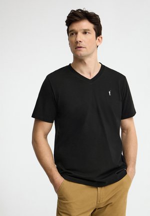 Mężczyzna o ciemnych włosach, ubrany w czarną koszulkę z dekoltem w serek i brązowe spodnie, stojący z rękami w kieszeniach, lekko patrzący na bok.