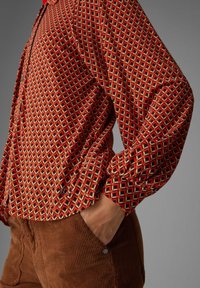 Rode blouse met geometrische patronen, voorzien van een ontspannen pasvorm en lange mouwen, gecombineerd met een paar bruine corduroy broeken met een zakdetail.