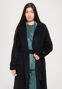 Melna vilnas tranšeja jaka ar jostu viduklī, izdurtu apkakli un pogām uz piedurknēm. Uzvilkta virs zila, ribota iekšējā slāņa.