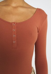 Rippierter langärmeliger Body in rostfarbener Optik mit einem weiten Rundhalsausschnitt und fünf metallenen Druckknöpfen in der Mitte. Weiche Textur.