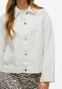 Vit denimjacka med krage, knäppning och stora framfickor. Tyget har en slät yta; ärmarna är något lösa.