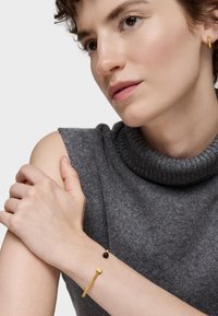 Złota bransoletka z teksturowanym paskiem i czarną koralik�ą, noszona na nadgarstku. Modelka w szarym swetrze z wysokim, prążkowanym kołnierzem.