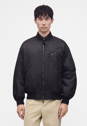 Jeune homme aux cheveux courts et noirs porte une veste bomber noire avec des poches zippées et un pantalon beige sur un fond blanc uni.
