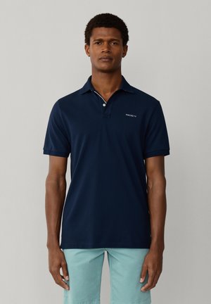 Mężczyzna ubrany w granatową koszulkę polo Hackett i jasnoniebieskie spodnie, stojący na tle gładkiego jasnoszarego tła.