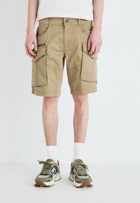 Khaki szorty cargo z wieloma bocznymi kieszeniami, paskami na pasek i zapięciem na guzik. Noszone z białymi skarpetkami i zielonymi sneakersami z akcentami.