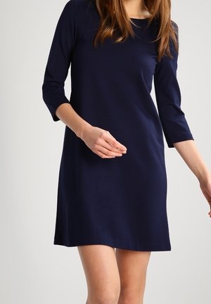 Nainen tummansininen polvipituinen mekko, jossa on kolmevartiset hihat, seisoo vaalean yksinkertaisen taustan edessä.