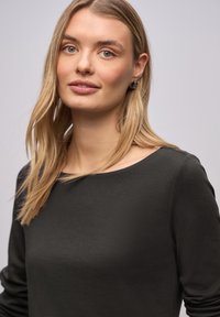 Czarna bluzka z długim rękawem i dekoltem w łódkę, wykonana z gładkiej tkaniny. Charakteryzuje się luźnym krojem i miękką fakturą, noszona przez modelkę z długimi blond włosami.