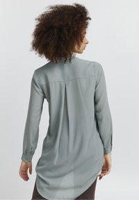 Light green long-sleeve blouse with a straight cut, pleated back, and buttoned cuffs; smooth, sheer fabric with a relaxed fit.