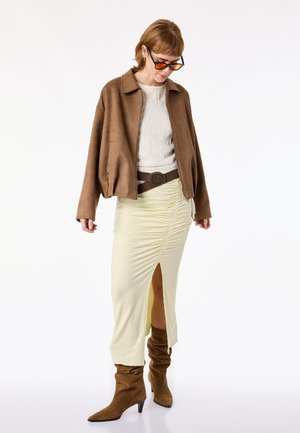 Kvinde iført brun ruskindjakke, hvid tekstureret top, cremefarvet rynket nederdel med slids foran, brun bælte, løse brune støvler og solbriller med orange skær.