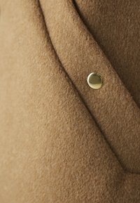 Tessuto in lana color beige con una superficie morbida e testurizzata, caratterizzato da un bottone dorato su una sezione ripiegata, che mette in risalto un design semplice.