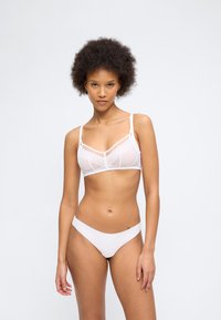 Νεαρή γυναίκα με αφρικανικό χτένισμα, που φοράει ένα λευκό δίχτυο μπραλέτο και ασορτί άνετα λευκά εσώρουχα, στέκεται μπροστά σε ένα απλό ανοιχτό υπόβαθρο.