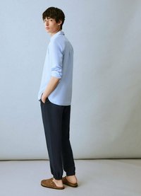 Camicia azzurra chiaro con maniche arrotolate, abbinata a pantaloni scuri con risvolto e scarpe slip-on marroni. Design minimalista e texture morbide.