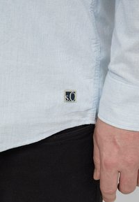 Hellblaues, gewebtes Hemd mit einem sichtbaren Etikett "s.O" am Saum, kombiniert mit schwarzen Stoffhosen. Das Hemd hat eine strukturierte Optik.