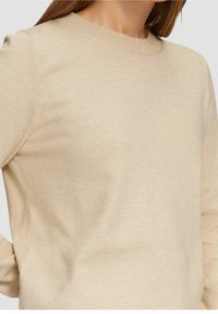 Nainen, jolla on yllään beige pitkähihainen pyöreäkauluksinen neule, kuvattuna hartioista vyötäröön yksiväristä taustaa vasten.