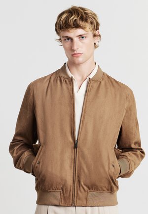 Giovane uomo che indossa una giacca bomber in suede marrone chiaro con polsini e colletto a coste, in piedi con le mani nelle tasche su uno sfondo neutro.