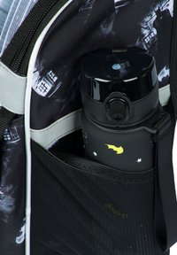 Bottiglia d'acqua nera con un logo a pipistrello giallo, riposta in una tasca a rete di uno zaino con un design a motivi e accenti grigi.