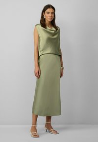 Olijfgroen satijnen jurk met een gedrapeerde mouwloze top en een rechte rok. Kenmerkt zich door een gladde textuur en een minimalistisch ontwerp. Beige hakken met bandjes.