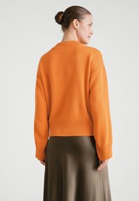 Dzianinowy pomarańczowy sweter z prążkowanym dołem i mankietami, o luźnym kroju i szerokich rękawach, zestawiony z jedwabistą brązową spódnicą.