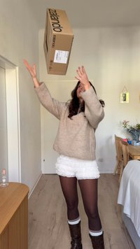 Kardboard kast, millel on kiri "Zalando", on õhus. Isik kannab beeži ülerööriga jakki, valgeid volangelehega lühikesi pükse, pruune saapaid ja mustriga sukkpükse.