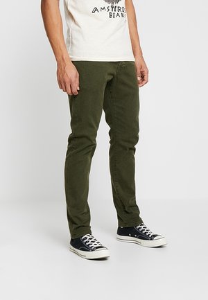 Homme portant un pantalon slim vert olive, des baskets montantes noires avec des lacets blancs, debout devant un fond clair uni.