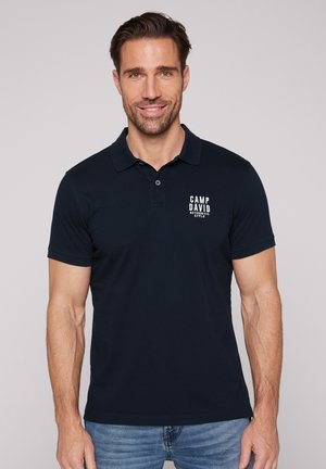 Lächelnder Mann, der ein schwarzes Poloshirt mit dem Text "Camp David Authentic Style" trägt, kombiniert mit blauen Jeans, auf einem einfarbigen hellen Hintergrund.