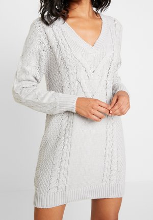 Femme portant une robe-pull en maille torsadée gris clair avec un col en V et des manches longues, les mains posées délicatement devant.