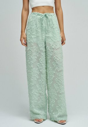 Mujer lleva pantalones anchos transparentes color verde menta con patrones florales texturizados y un top corto blanco, posando frente a un fondo liso.