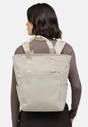 Beige corduroy backpack with dual short handles and adjustable straps. Front logo and side pockets for additional storage.
