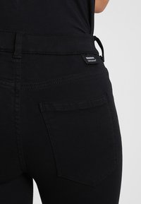 Jeans negros de talle alto con un corte ajustado. Incluyen bolsillos traseros, costuras sutiles y una etiqueta cerca de la cintura. Textura suave.
