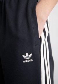 Svarta träningsbyxor med vita sidostråk, med en broderad adidas-logotyp på framsidan av fickområdet. Mjuk, bomullsblandad tyg.