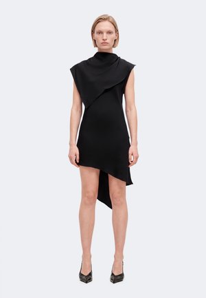 Le mannequin se tient devant un fond clair uni, vêtu d’une robe noire sans manches au décolleté et à l’ourlet drapés de façon asymétrique, ainsi que de chaussures noires à talons.