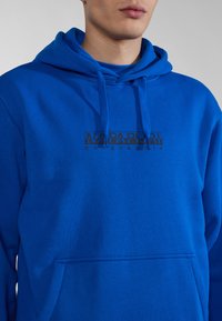 Sudadera con capucha azul hecha de una tela suave, con un bolsillo frontal y un diseño de logo impreso en negro en el pecho. Capucha ajustable con cordón.