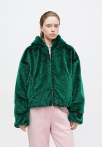 Grüne Kunstfell-Zip-Jacke mit Kapuze, die eine weiche Textur und lange Ärmel aufweist. Über hellrosa Hosen getragen, mit einem kontrastierenden Detail an den Bündchen.