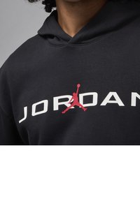 Primo piano di una persona che indossa un hoodie nero con il testo bianco "JORDAN" e il logo Jumpman rosso centrato sul petto.
