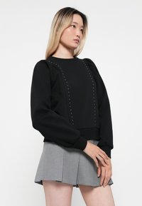 Sweatshirt noir avec des manches bouffantes, des détails cloutés sur le devant et une taille côtelée, associé à une jupe plissée grise.