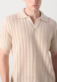Beige knitted polo shirt with short sleeves, featuring a textured, open weave pattern and a contrasting collar.
