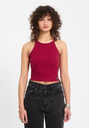 Jeune femme aux cheveux bouclés portant un crop top bordeaux sans manches et côtelé, ainsi qu'un jean noir taille haute, debout devant un fond blanc.