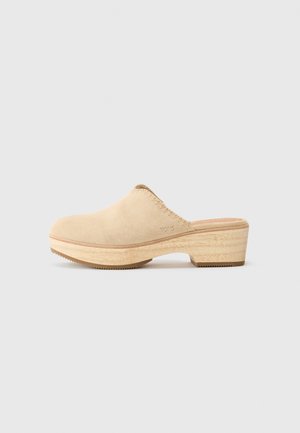 Beige Wildleder-Clogs mit einer Holzplattform und abgerundetem Zeh, sowie gesteppten Details entlang des Randes. Strukturiertes Profil für besseren Halt.