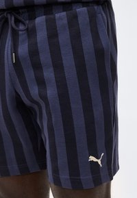 Κάτω μέρος σορτς με κάθετες ρίγες σε ναυτικό μπλε και μαύρο χρώμα, με κορδόνι στη μέση και μικρό λευκό λογότυπο Puma κοντά στο τελείωμα στο δεξί πόδι.