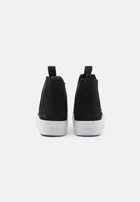 Svarta mockastövlar med vita gummisulor. Har en dragtab för enkel påtagning och en minimalistisk design, sedda bakifrån.