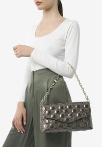 Metallisch grijze quilted handtas met kettinghengsel, voorzien van een klep sluiting en een geperst bloemenpatroon; vastgehouden door een model in een witte top.