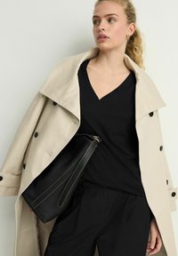 Beige trenchcoat met zwarte knopen, gedragen over een zwart V-hals top. Een zwarte leren tas met een gouden accent en lichte stiksels wordt vastgehouden.