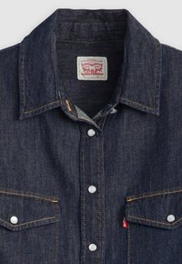 Camisa de denim azul escuro com colarinho pontudo, dois bolsos no peito e botões de pressão brancos. Apresenta costuras laranjas e uma etiqueta de marca no interior.