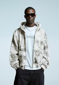 Capuchon zip-up jas in crème met bruine abstracte patronen, gelaagd over een wit t-shirt met zwarte tekst. Zwarte zonnebril en kettingen worden gedragen.