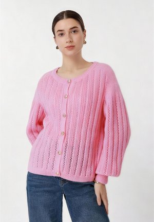 Ženska s svetlo poltjo in temnimi lasmi nosi roza pleteno jopico z gumbi in napihnjenimi rokavi ter modre kavbojke, stoji ob belem ozadju.