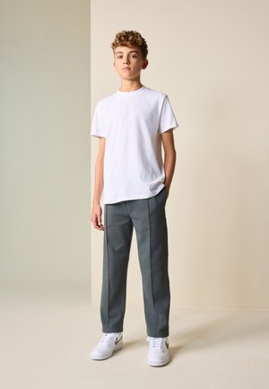 Adolescent debout avec les mains dans les poches, portant un t-shirt blanc, un pantalon gris et des baskets blanches sur un fond beige bicolore.