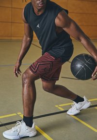 Camiseta sin mangas negra, pantalones cortos burdeos con patrón geométrico negro, zapatillas deportivas blancas, driblando un balón de baloncesto en una cancha.