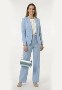 Femme souriante, portant un tailleur-pantalon bleu clair avec un chemisier blanc, tenant un sac à main argenté métallique et des talons hauts argentés à brides.